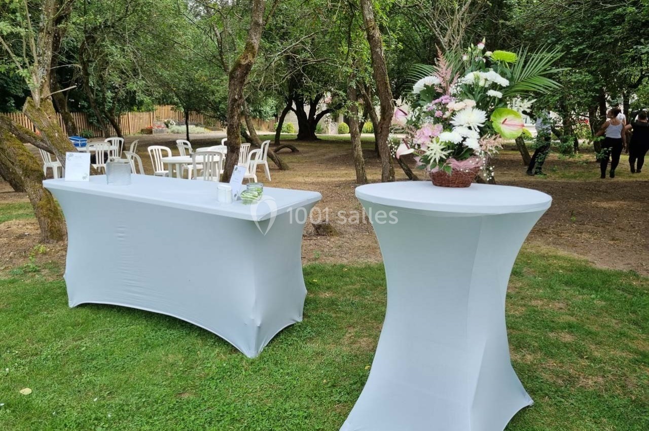 Table dressée avec nappe blanche et décor floral, installée en extérieur sur une pelouse entourée d'arbres.