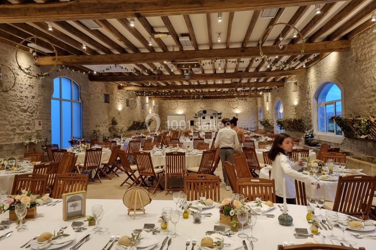 Salle de réception en pierre avec tables dressées pour un événement, décorée de fleurs et éclairée par des lumières…