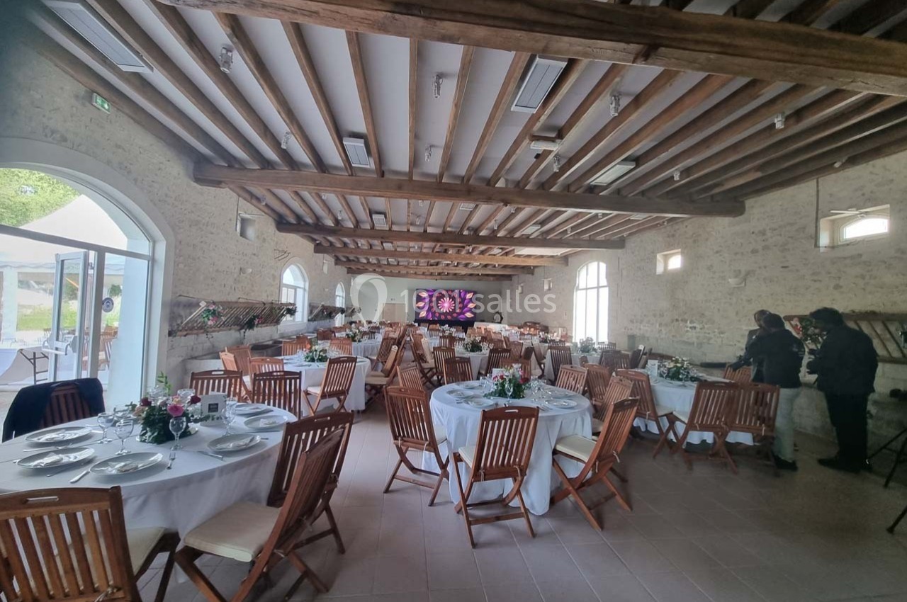 Salle de réception avec tables rondes dressées, chaises en bois, poutres apparentes et écran au fond.