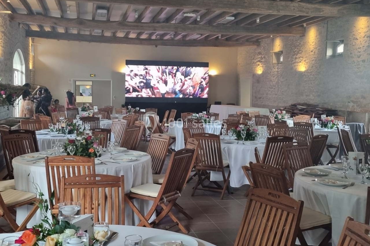 Salle de réception avec tables rondes dressées, chaises en bois et écran géant affichant une scène festive.