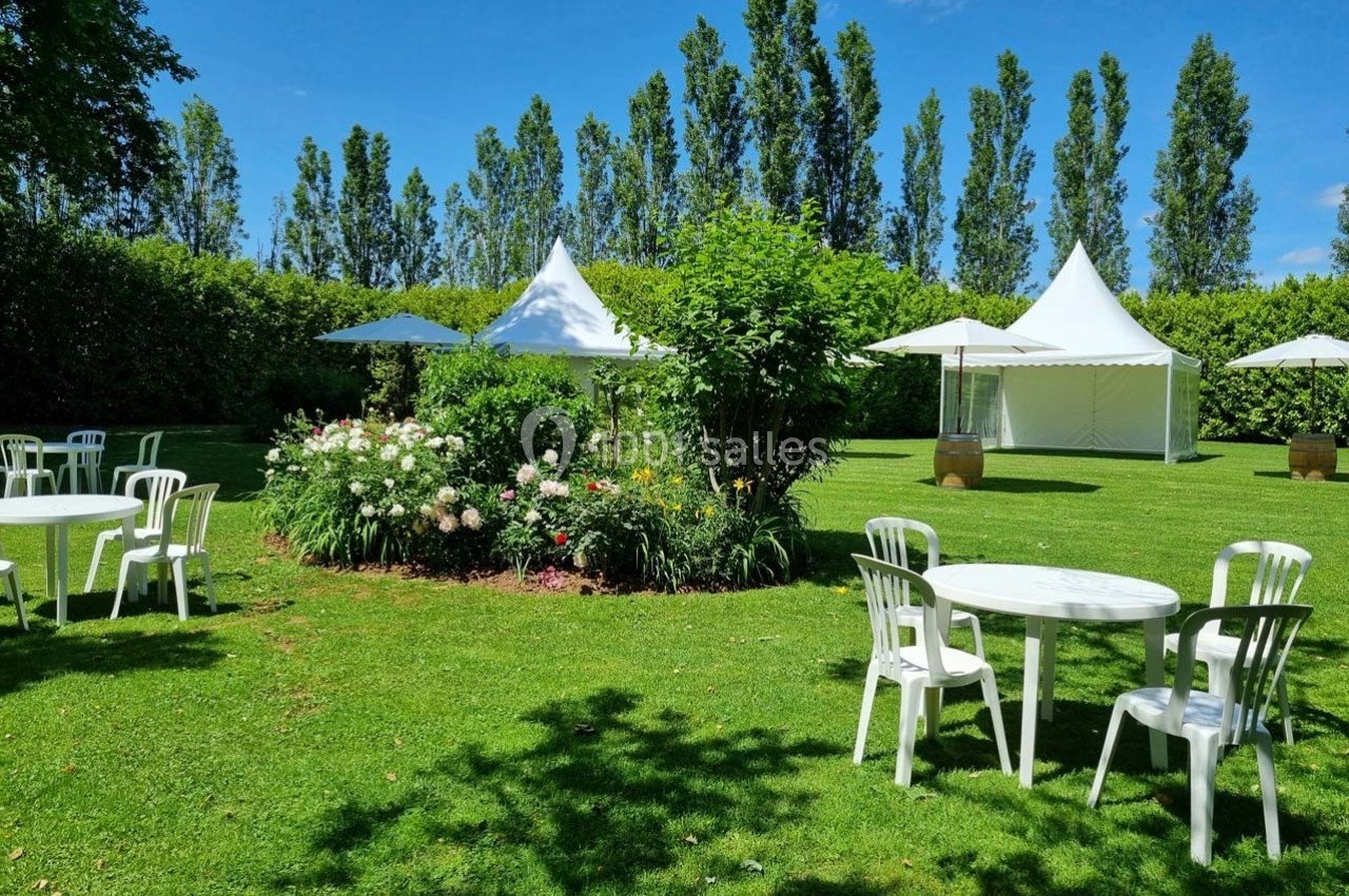 Jardin verdoyant avec tables blanches, chaises et tentes installées pour un événement en plein air.
