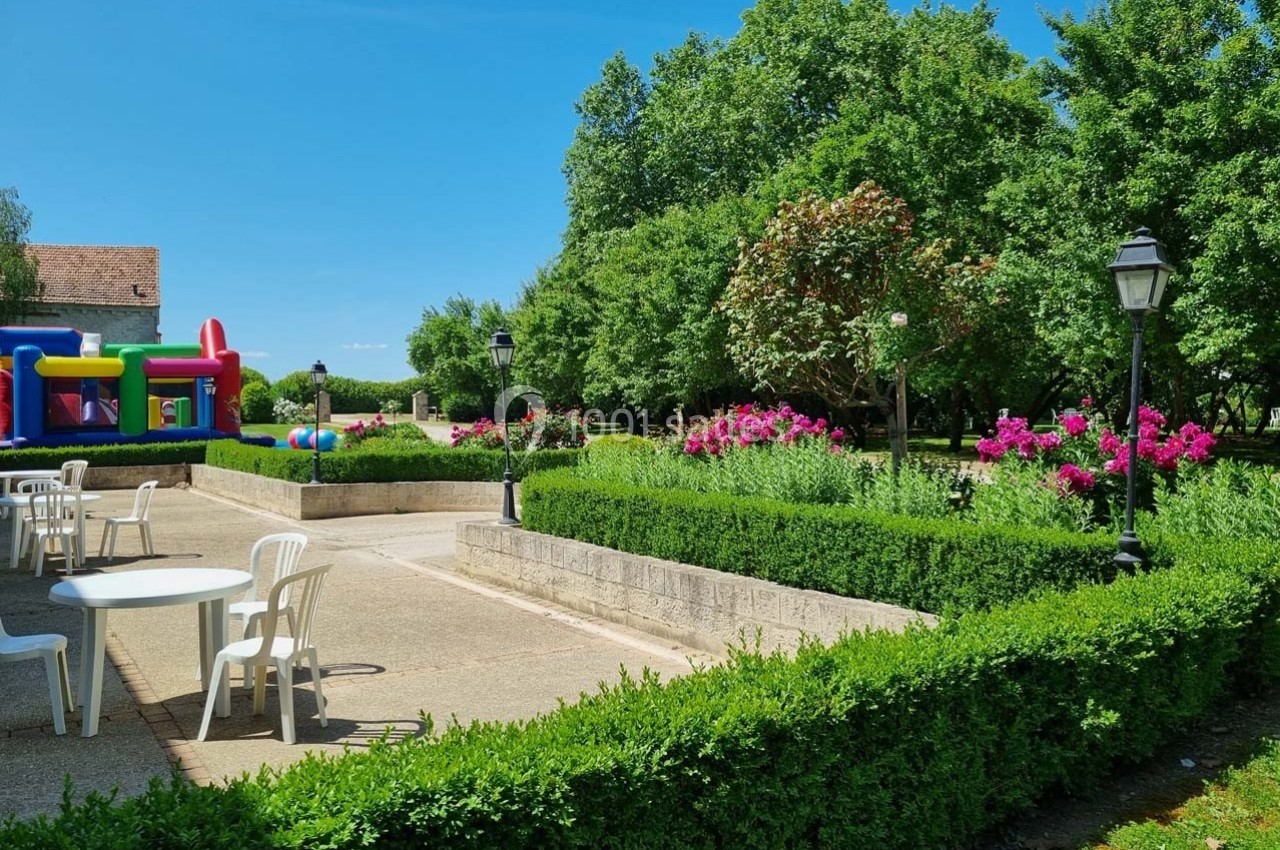 Jardin avec haies taillées, fleurs roses, tables blanches et structures gonflables colorées sous un ciel bleu.