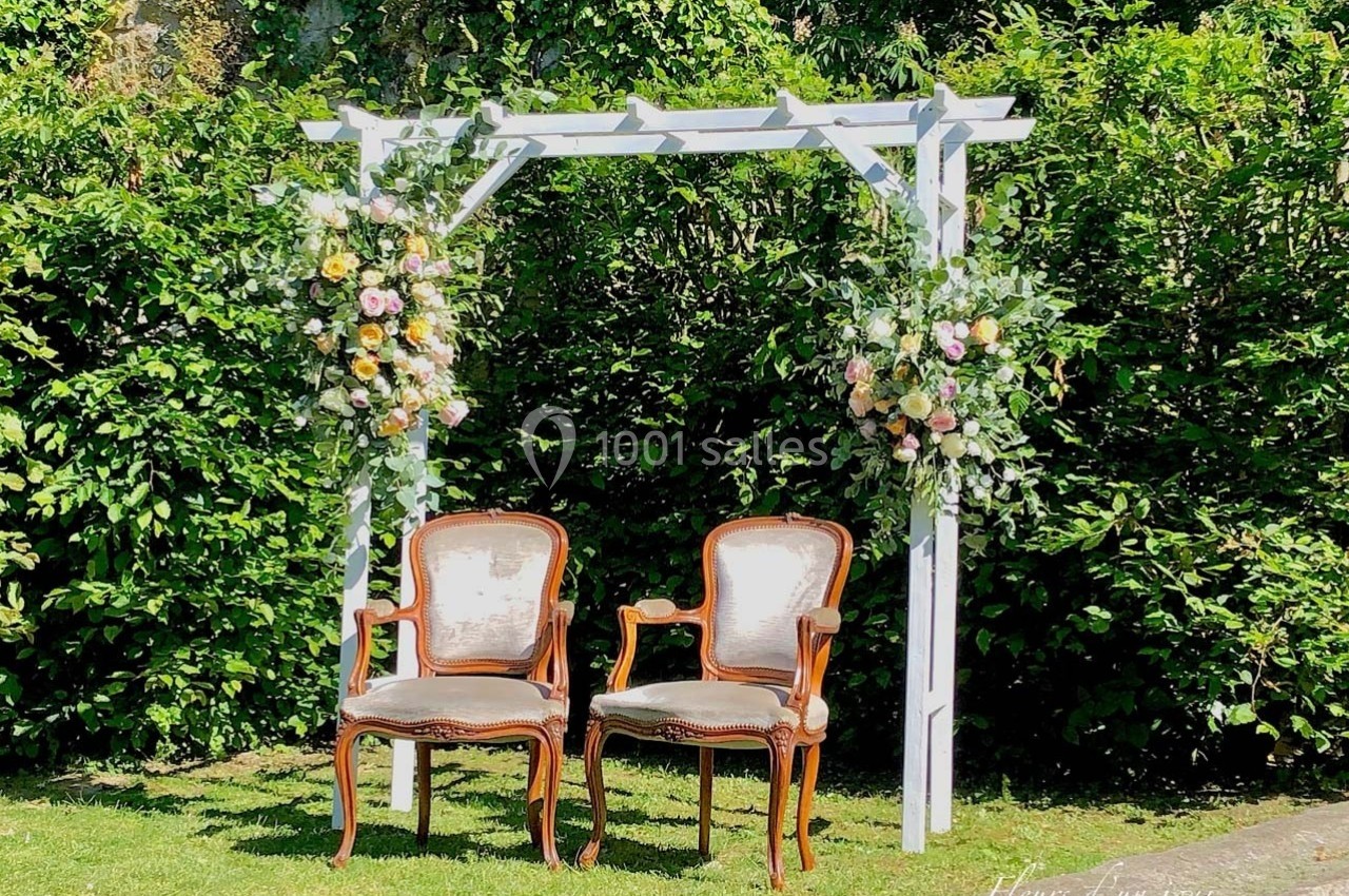 Deux chaises anciennes sous une arche blanche décorée de fleurs, placées dans un jardin verdoyant.