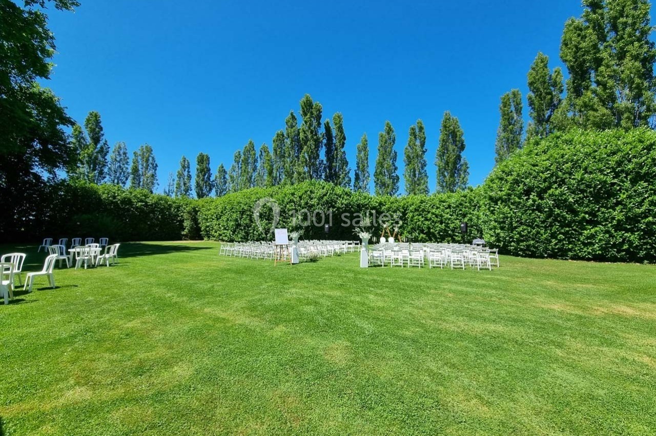 Chaises blanches disposées sur une pelouse verte entourée de haies et d'arbres sous un ciel bleu.