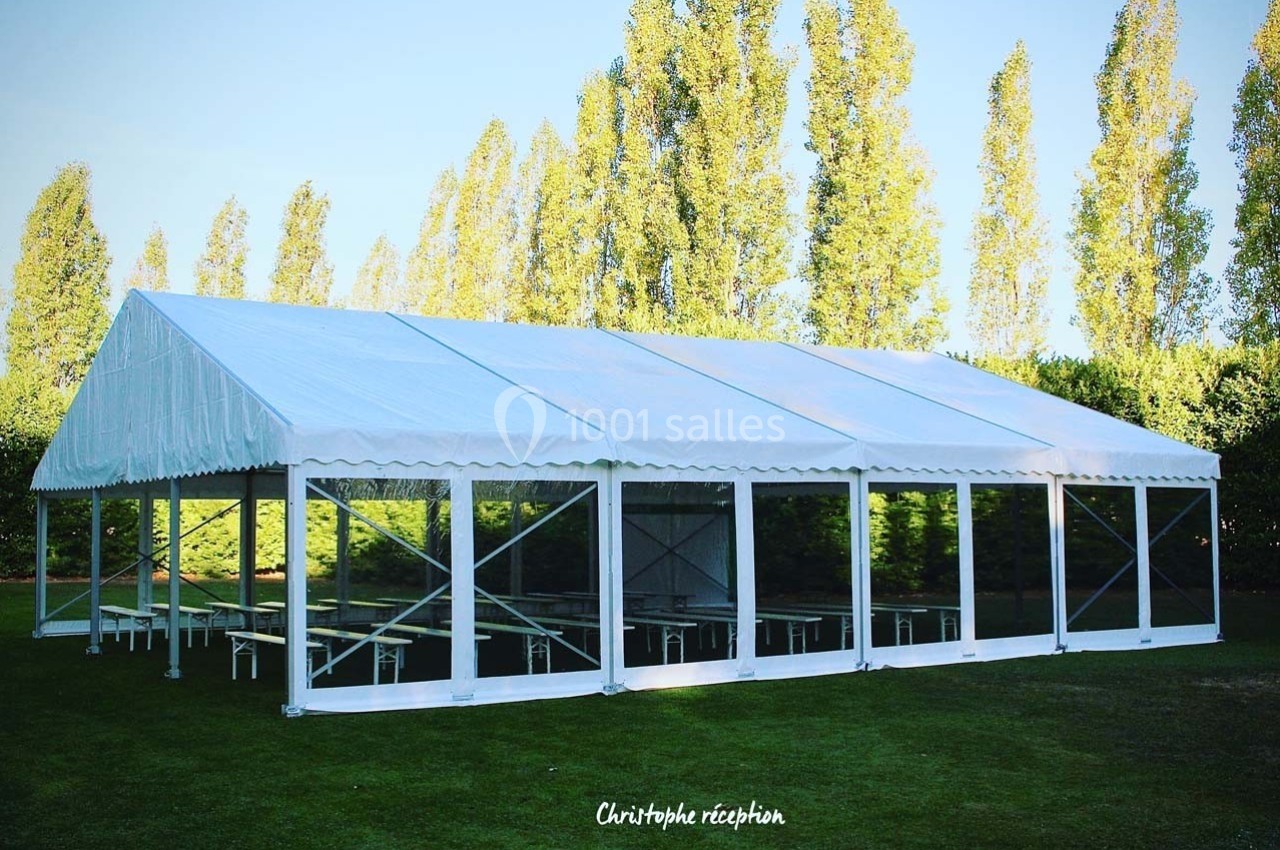 Grande tente blanche installée sur une pelouse, entourée d'arbres, avec des parois transparentes.