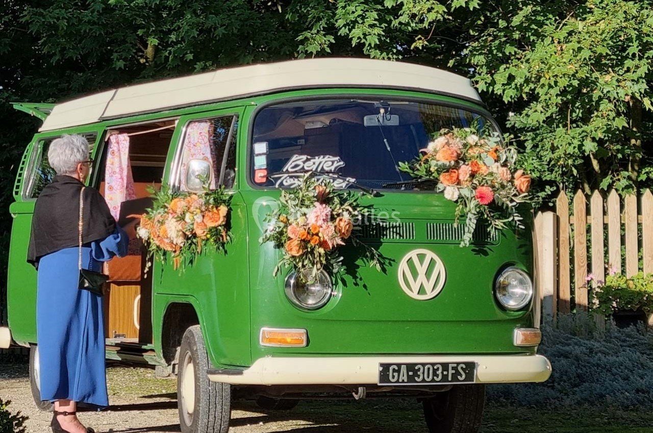 Une femme près d'un van Volkswagen vert décoré de fleurs, stationné devant des arbres et une clôture en bois.