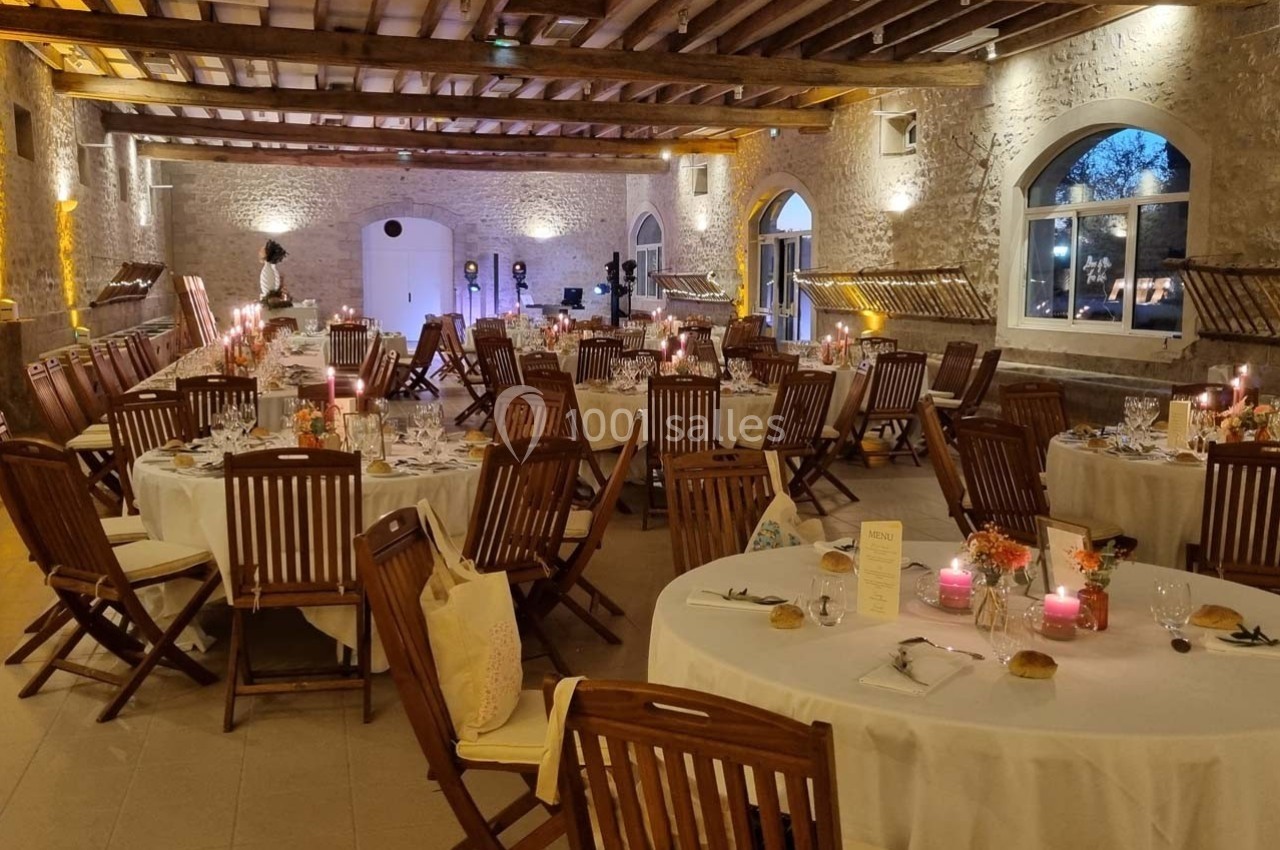 Salle de réception décorée avec des tables rondes dressées, chaises en bois et éclairage chaleureux.