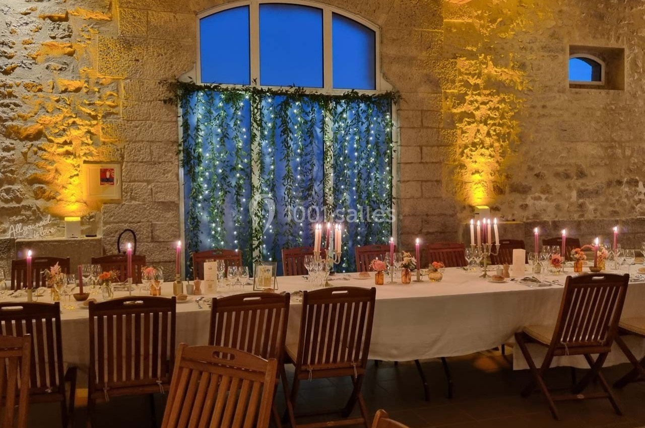 Grande table dressée pour un dîner, décorée de chandeliers et guirlandes lumineuses, dans une salle en pierre éclairée.