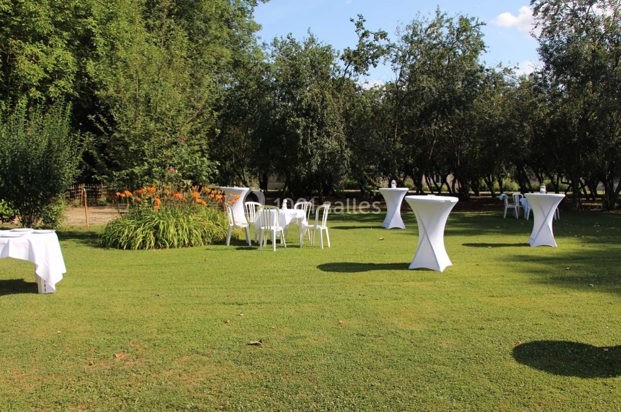 Tables hautes et chaises blanches disposées sur une pelouse verdoyante entourée d'arbres et de buissons.