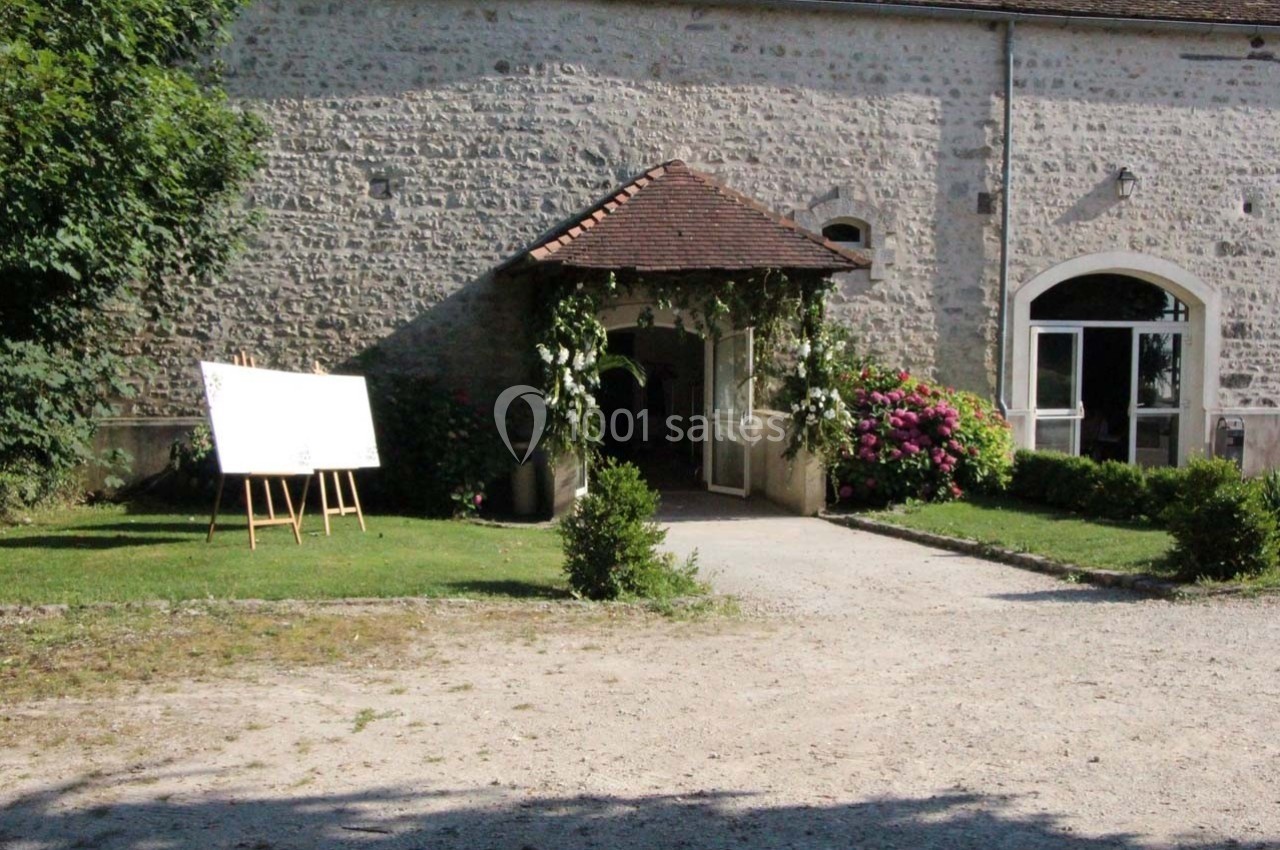 Entrée d'un bâtiment en pierre avec un porche fleuri, un tableau sur chevalet et des buissons autour.