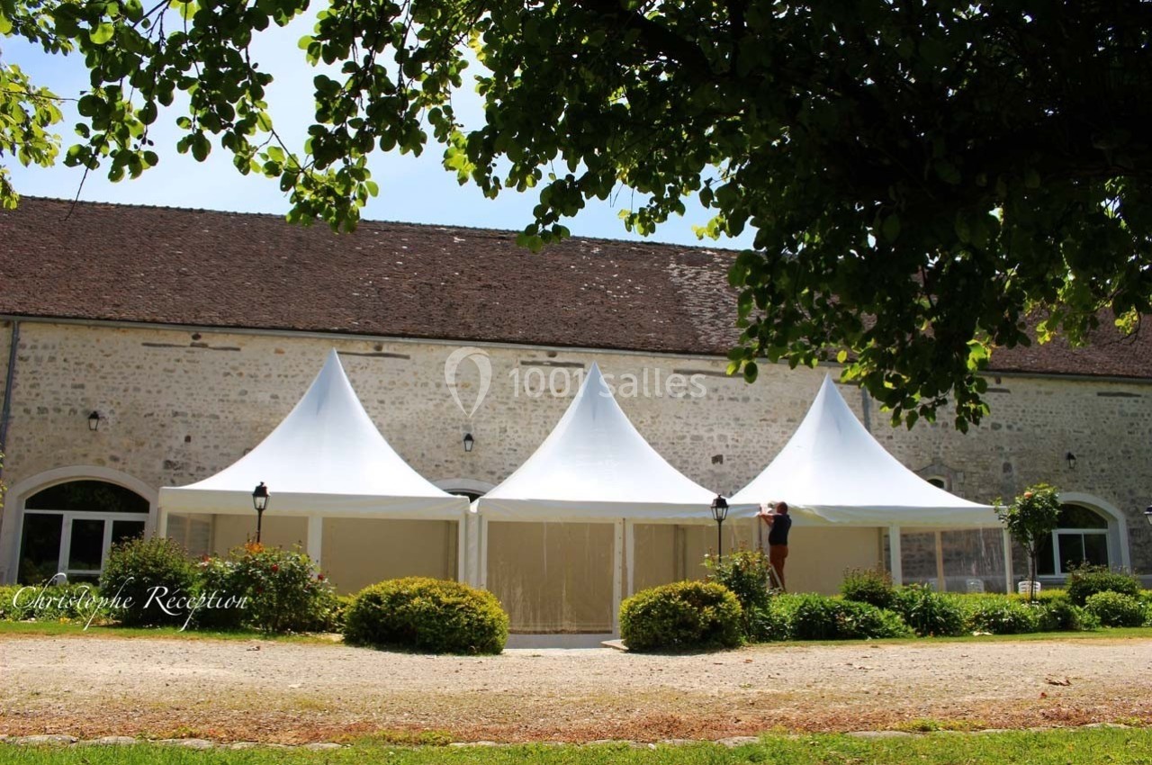 Trois tentes blanches installées devant un bâtiment en pierre, entourées de verdure et d'arbustes.