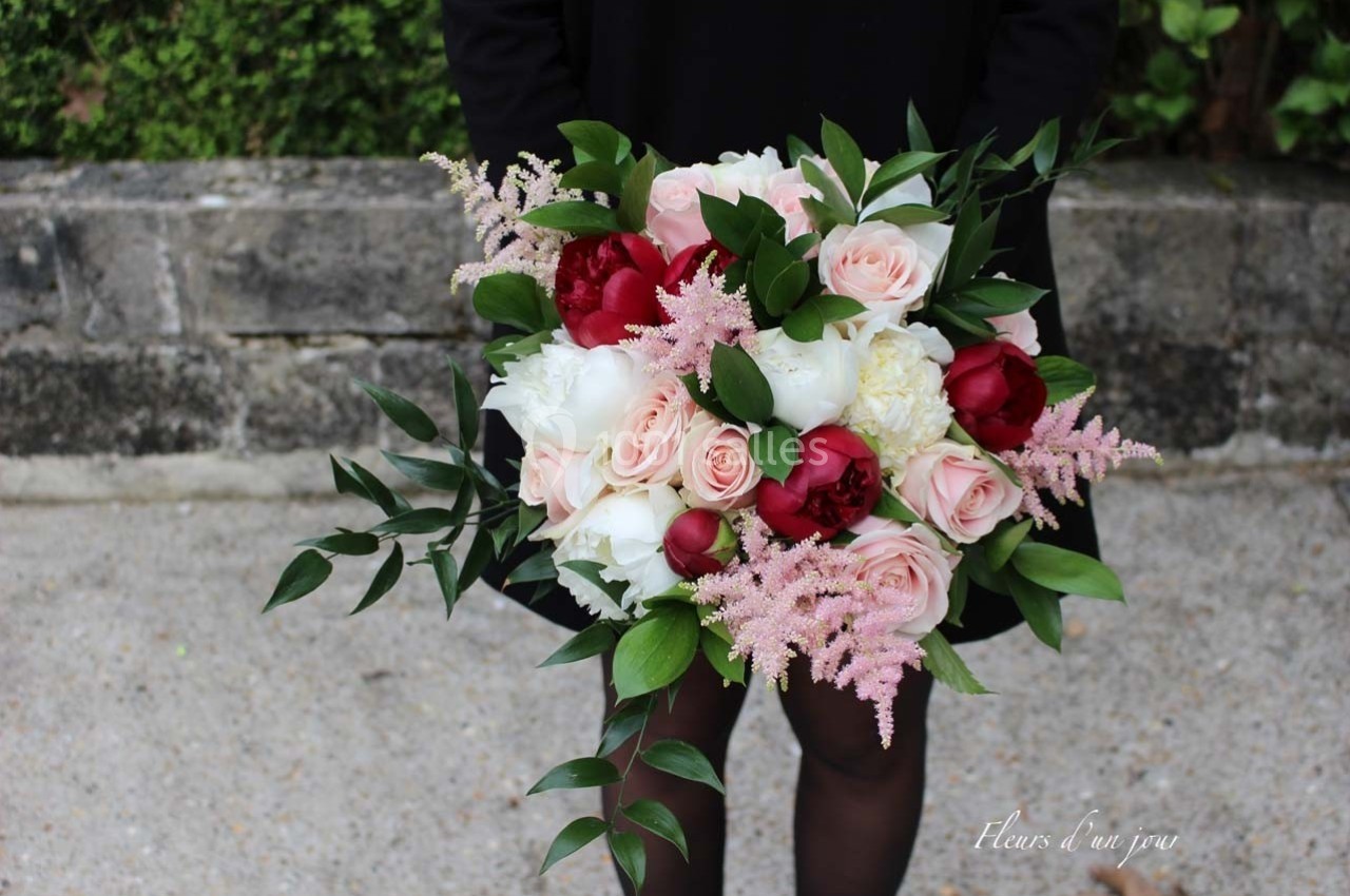 Bouquet de fleurs composé de roses, pivoines, astilbes roses et feuillage, tenu par une personne en arrière-plan flou.