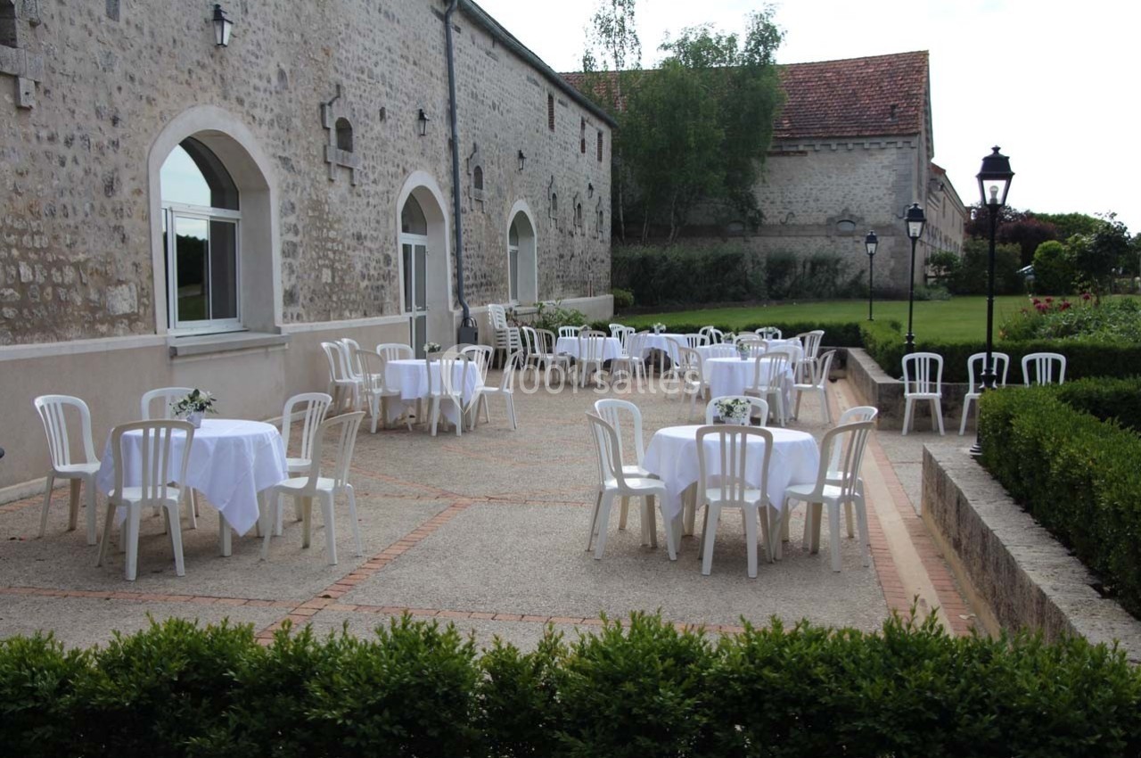 Terrasse en plein air avec des tables rondes dressées de nappes blanches, entourée de chaises en plastique blanc.