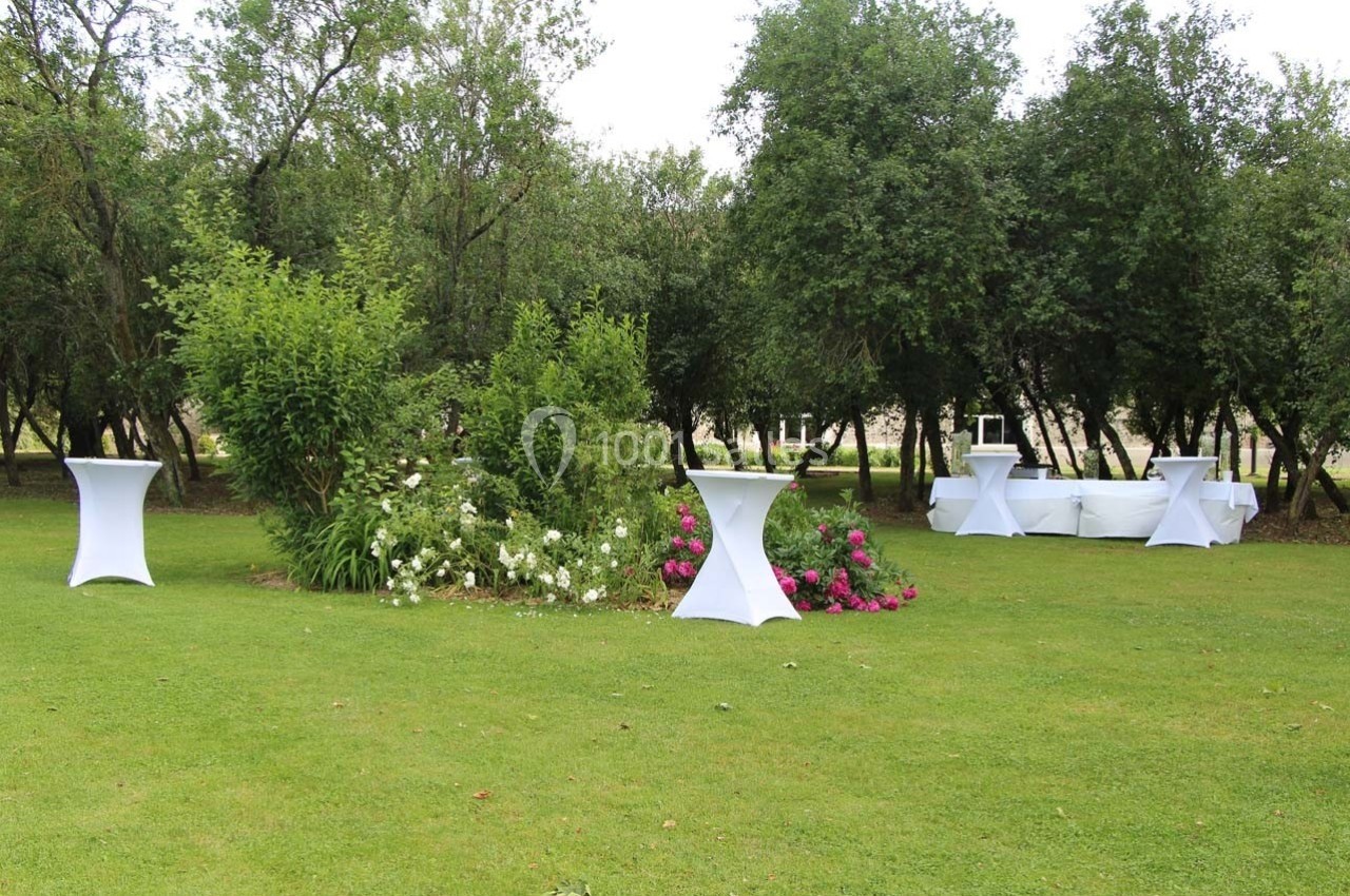 Jardin avec pelouse, tables hautes blanches et parterre de fleurs colorées entouré d'arbres.
