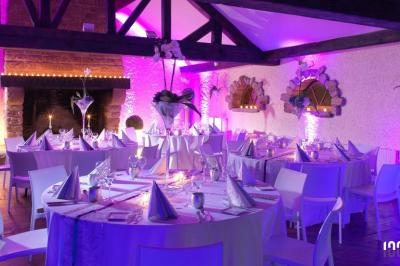 Salle de réception décorée avec des tables rondes, nappes blanches, éclairage violet et poutres apparentes.