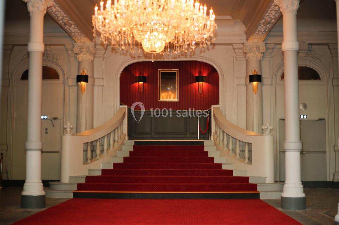 Escalier central recouvert de tapis rouge, encadré de balustrades blanches, sous un grand lustre en cristal.