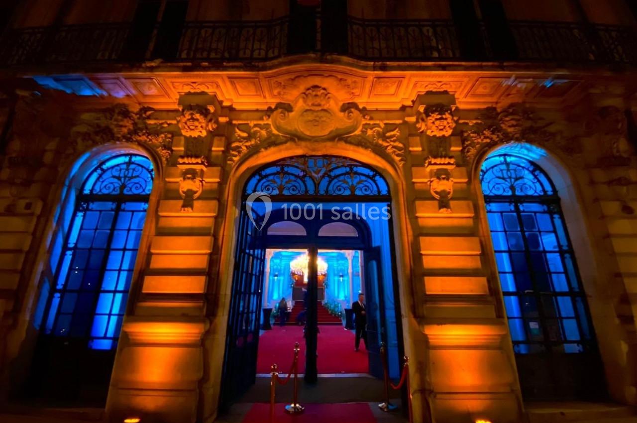 Façade d'un bâtiment illuminé en orange et bleu, avec une entrée ouverte sur un tapis rouge.