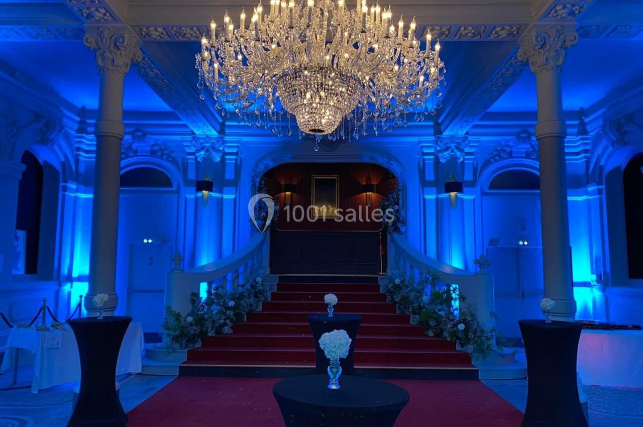 Salle élégante avec escalier central, tapis rouge, lustre imposant et éclairage bleu créant une ambiance raffinée.