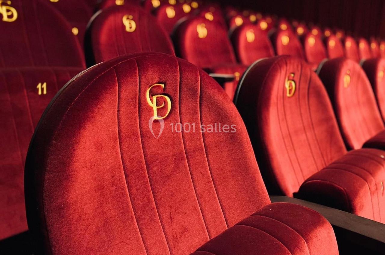 Rangée de fauteuils rouges en velours avec accoudoirs noirs et logo doré, dans une salle de spectacle ou de cinéma.