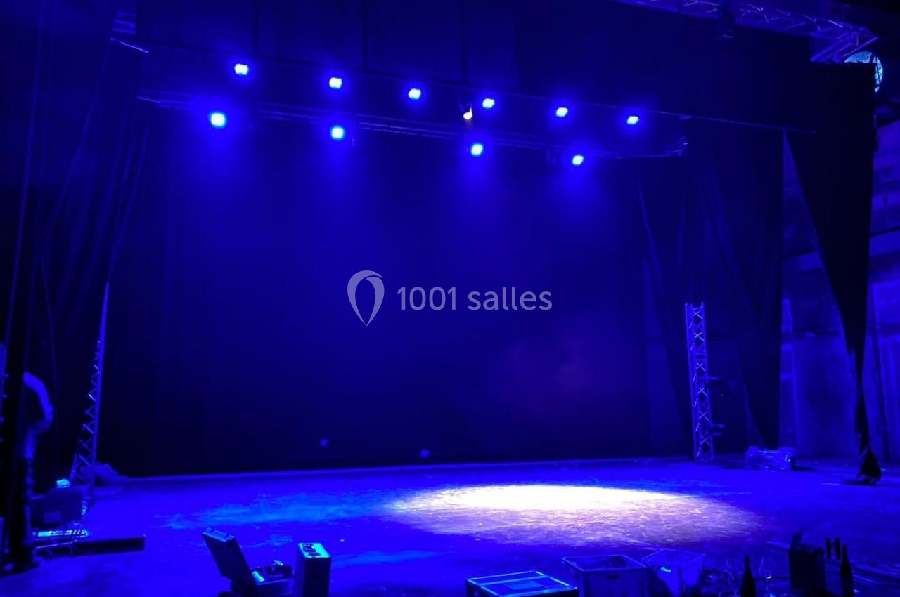Scène de théâtre vide éclairée par des projecteurs bleus, avec des équipements techniques au sol.