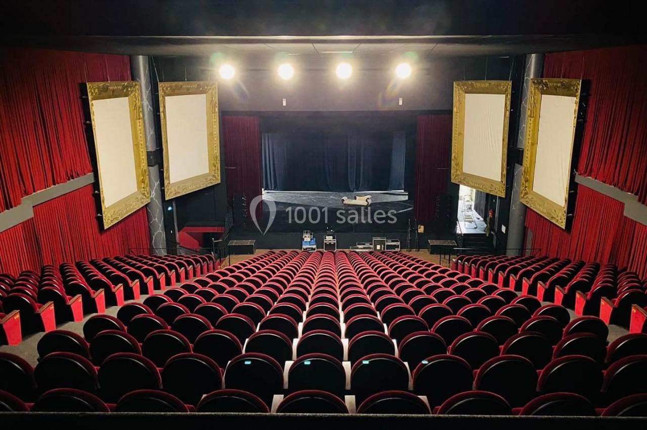 Salle de théâtre vide avec des rangées de sièges rouges, une scène éclairée et des rideaux rouges encadrant l'espace.