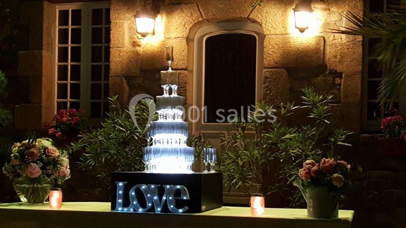 Centre de table lumineux avec une fontaine à champagne, entourée de fleurs, devant une façade en pierre éclairée.