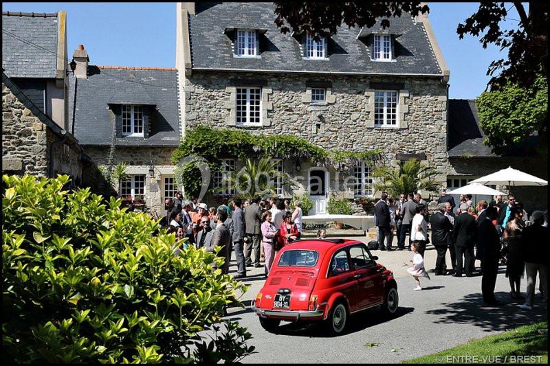 Une voiture rouge garée devant une maison en pierre, entourée de personnes lors d'un rassemblement en extérieur.