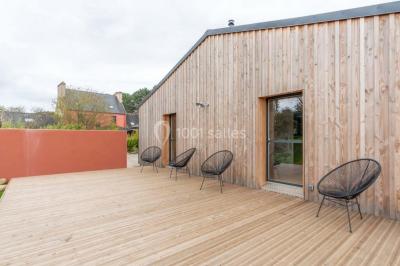 Terrasse en bois avec des chaises noires, adossée à une maison en bardage bois, sous un ciel nuageux.