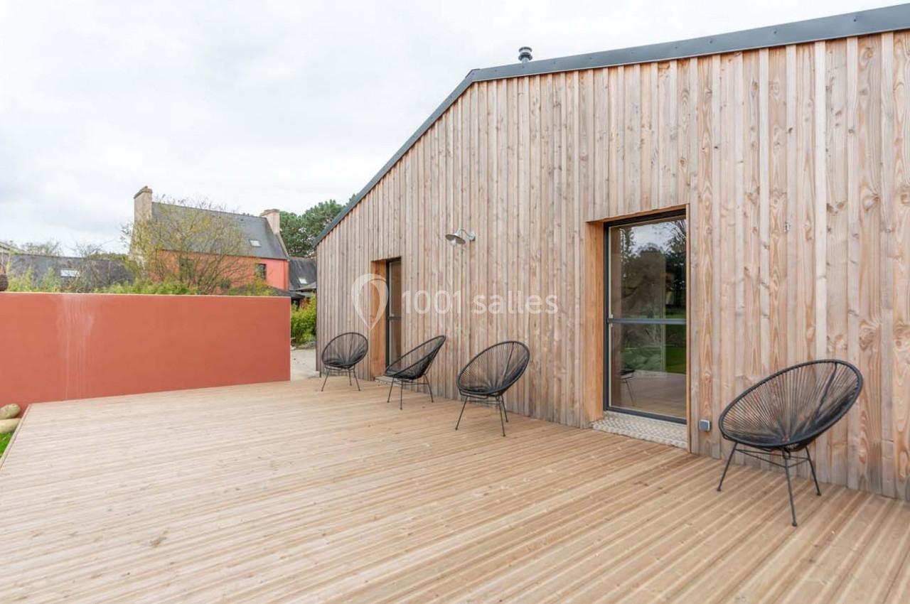 Terrasse en bois avec des chaises noires, adossée à une maison en bardage bois, sous un ciel nuageux.