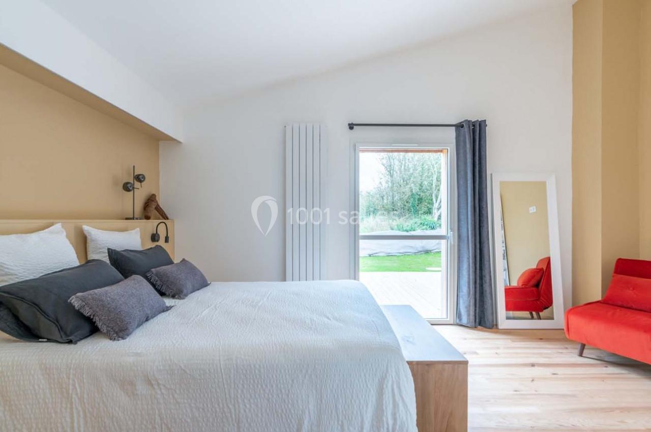 Chambre lumineuse avec lit double, coussins gris, miroir sur pied, fauteuil rouge et vue sur jardin par une porte vitrée.