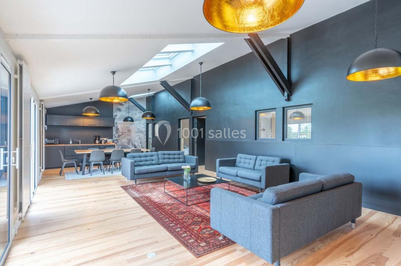 Espace de vie lumineux avec canapés gris, tapis rouge, cuisine ouverte et grandes suspensions dorées.