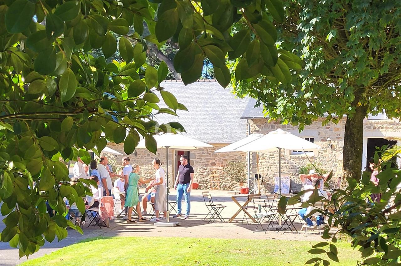 Des personnes discutent et se rassemblent autour de tables en extérieur, sous des arbres et près d'un bâtiment en pierre.