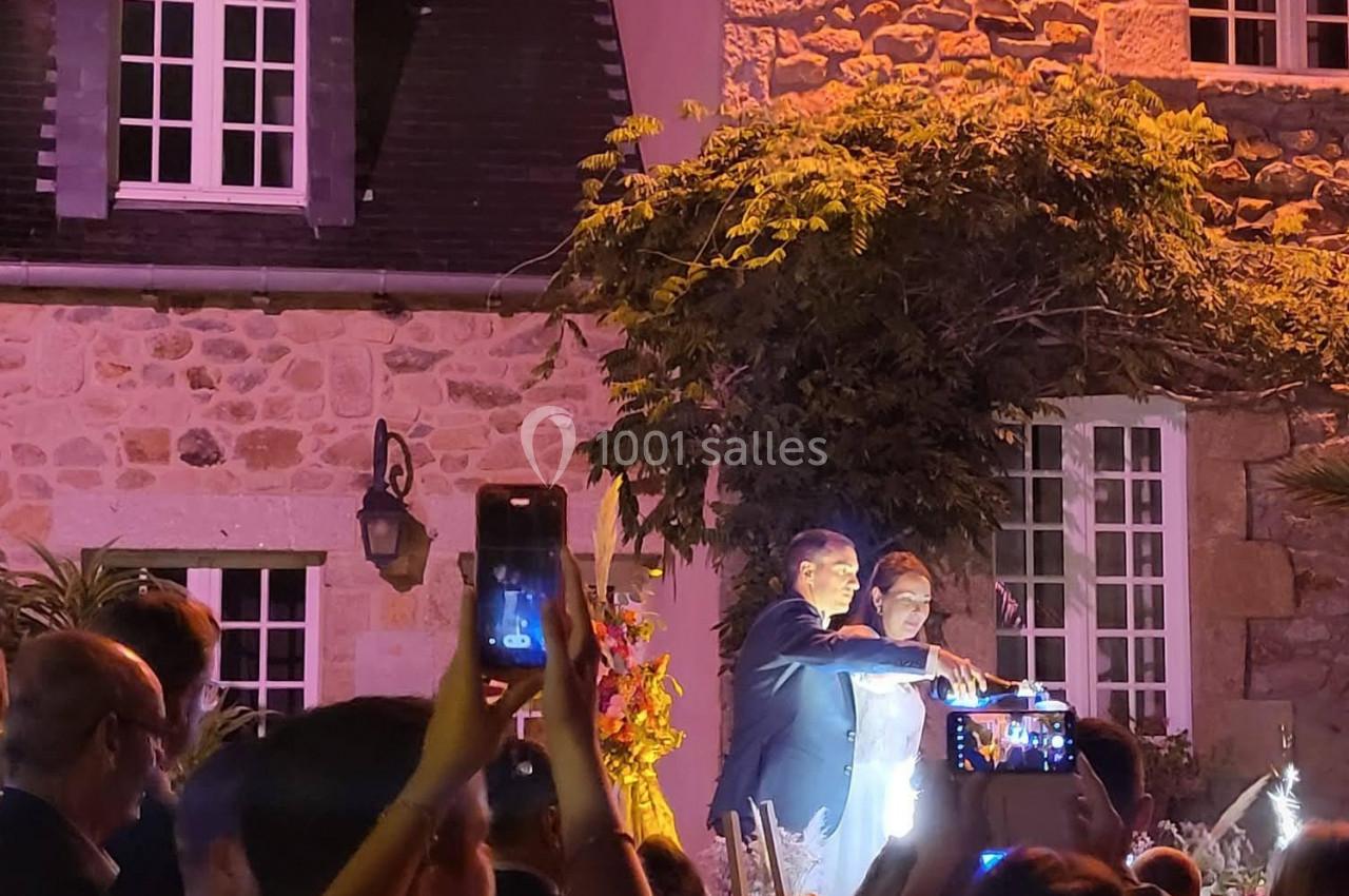 Un couple pose sous un éclairage nocturne devant une maison en pierre, entouré de personnes prenant des photos.