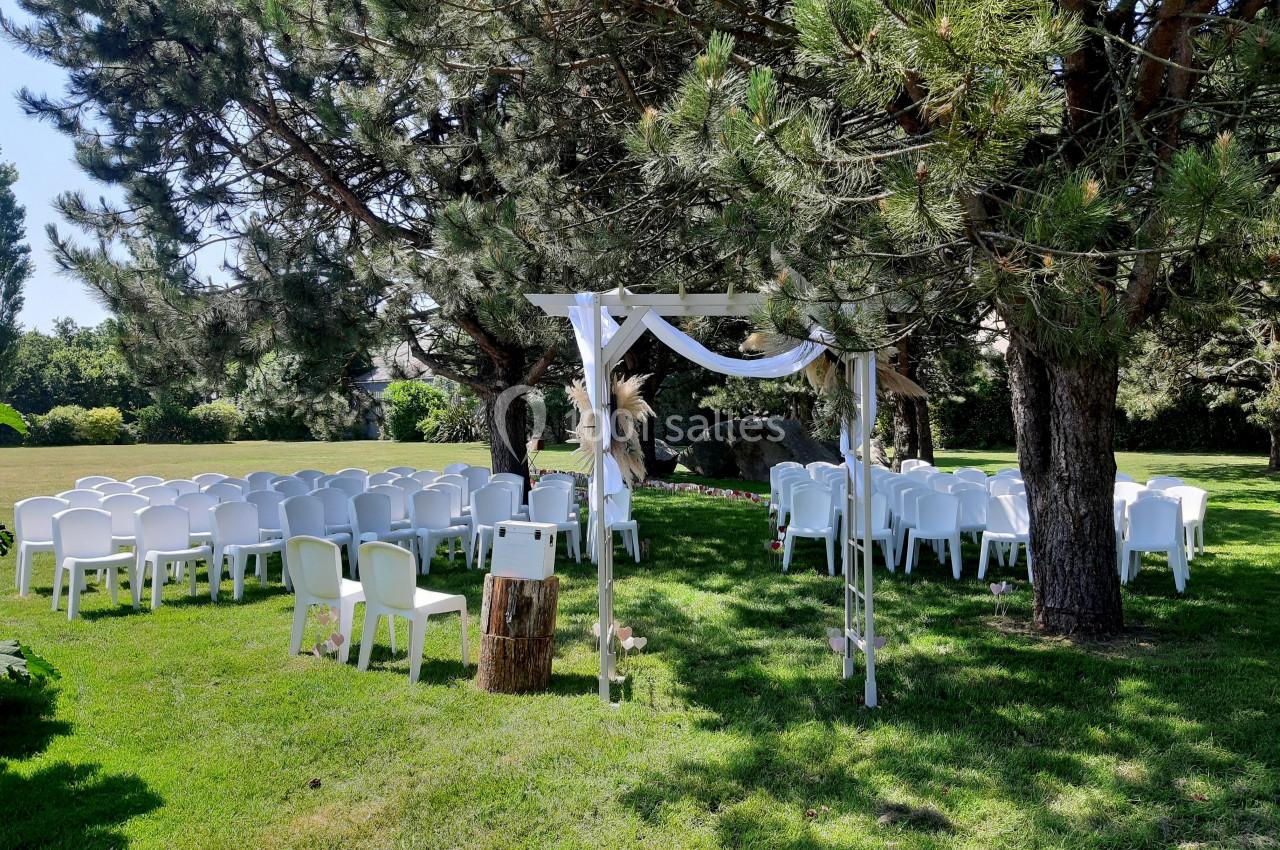 Chaises blanches disposées en extérieur sous des arbres, autour d'une arche décorée pour une cérémonie en plein air.
