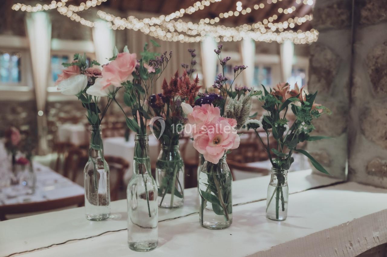 Bouquets de fleurs variées disposés dans des bouteilles en verre sur une table, avec des lumières suspendues en arrière-plan.