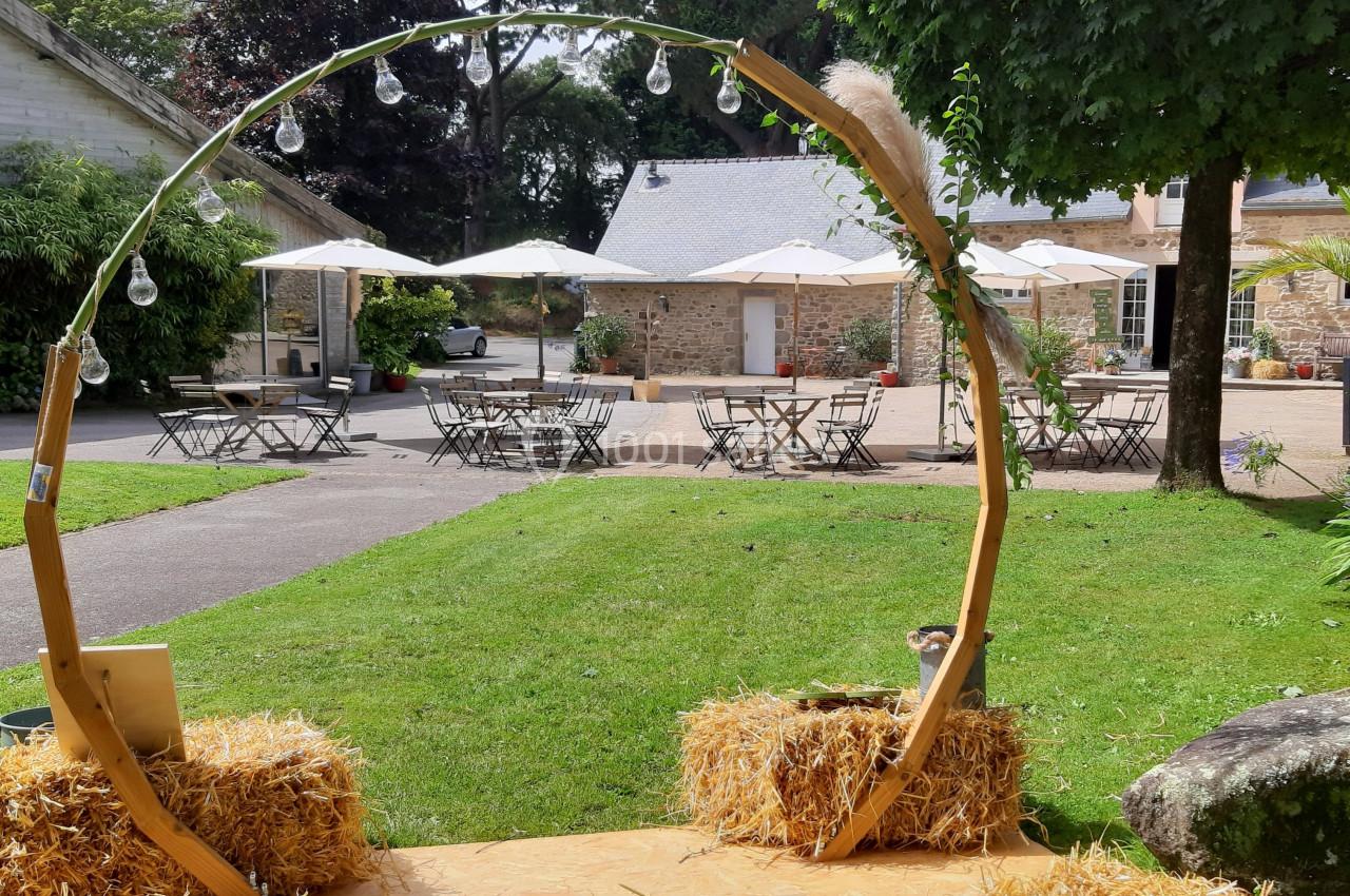 Arc décoratif en bois entouré de bottes de paille sur une pelouse, avec des tables et parasols en arrière-plan.