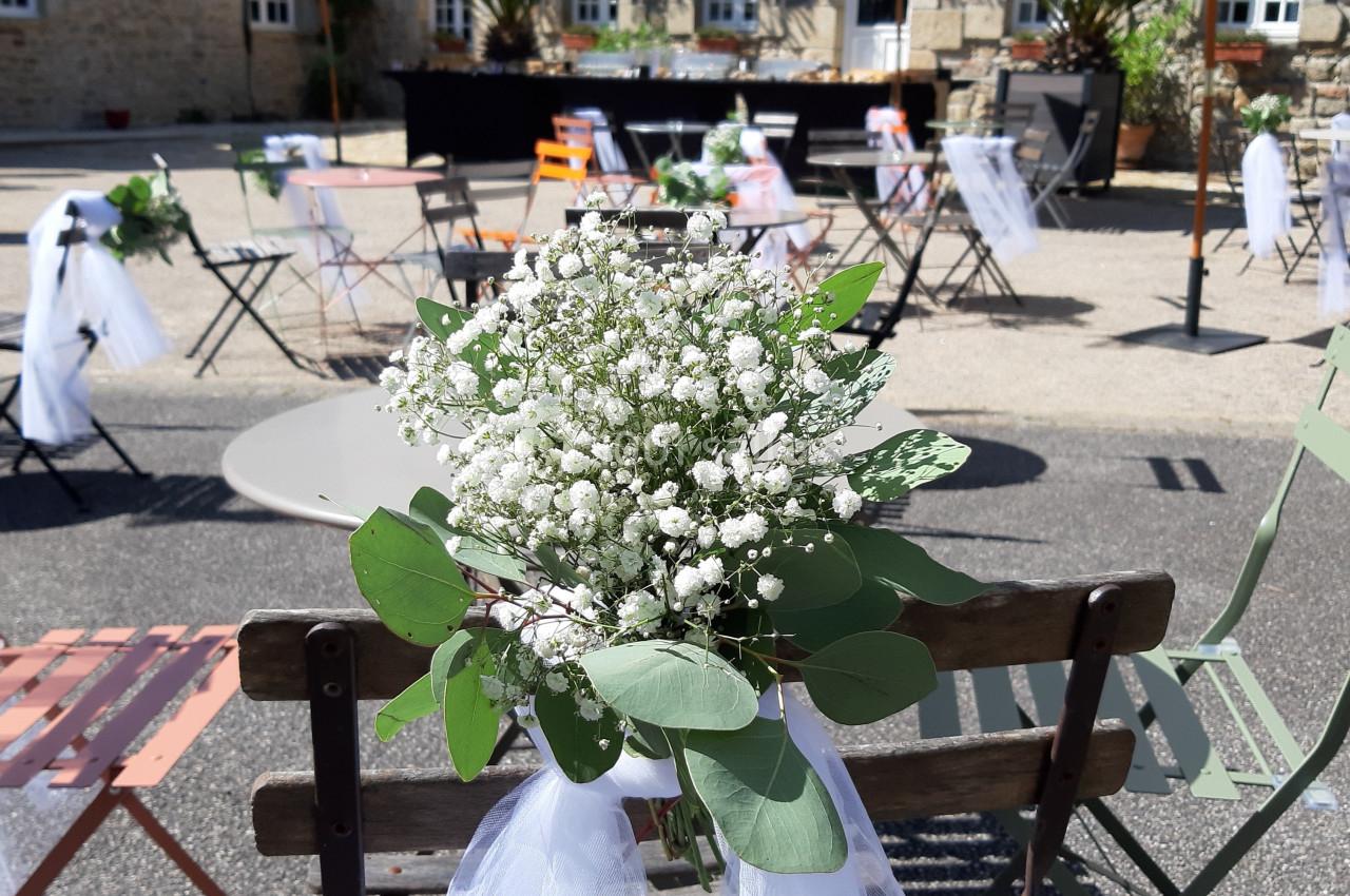 Chaises décorées de gypsophiles et de voiles blanches dans une cour ensoleillée devant un bâtiment en pierre.