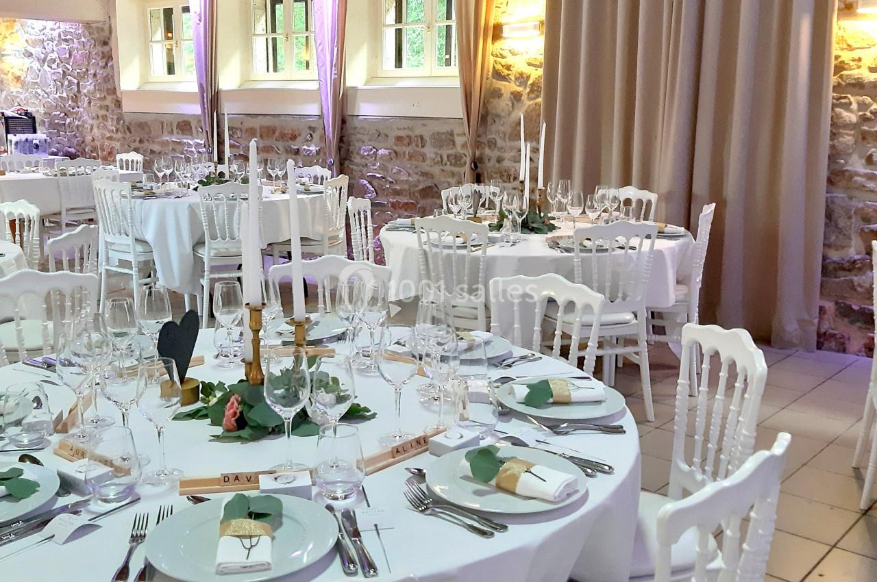 Salle de réception décorée avec des tables rondes dressées, nappes blanches, chandeliers et murs en pierre apparente.