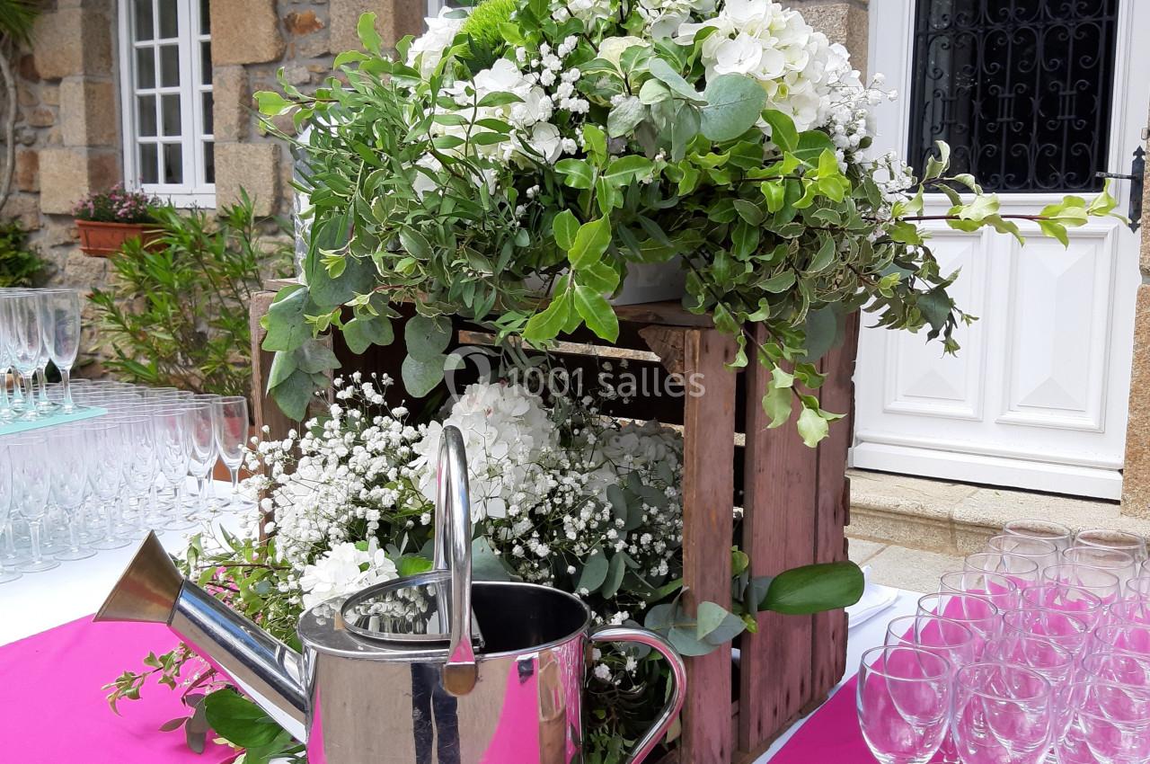 Arrangement floral avec des fleurs blanches et feuillages dans une caisse en bois, arrosoir en métal et verres en arrière…
