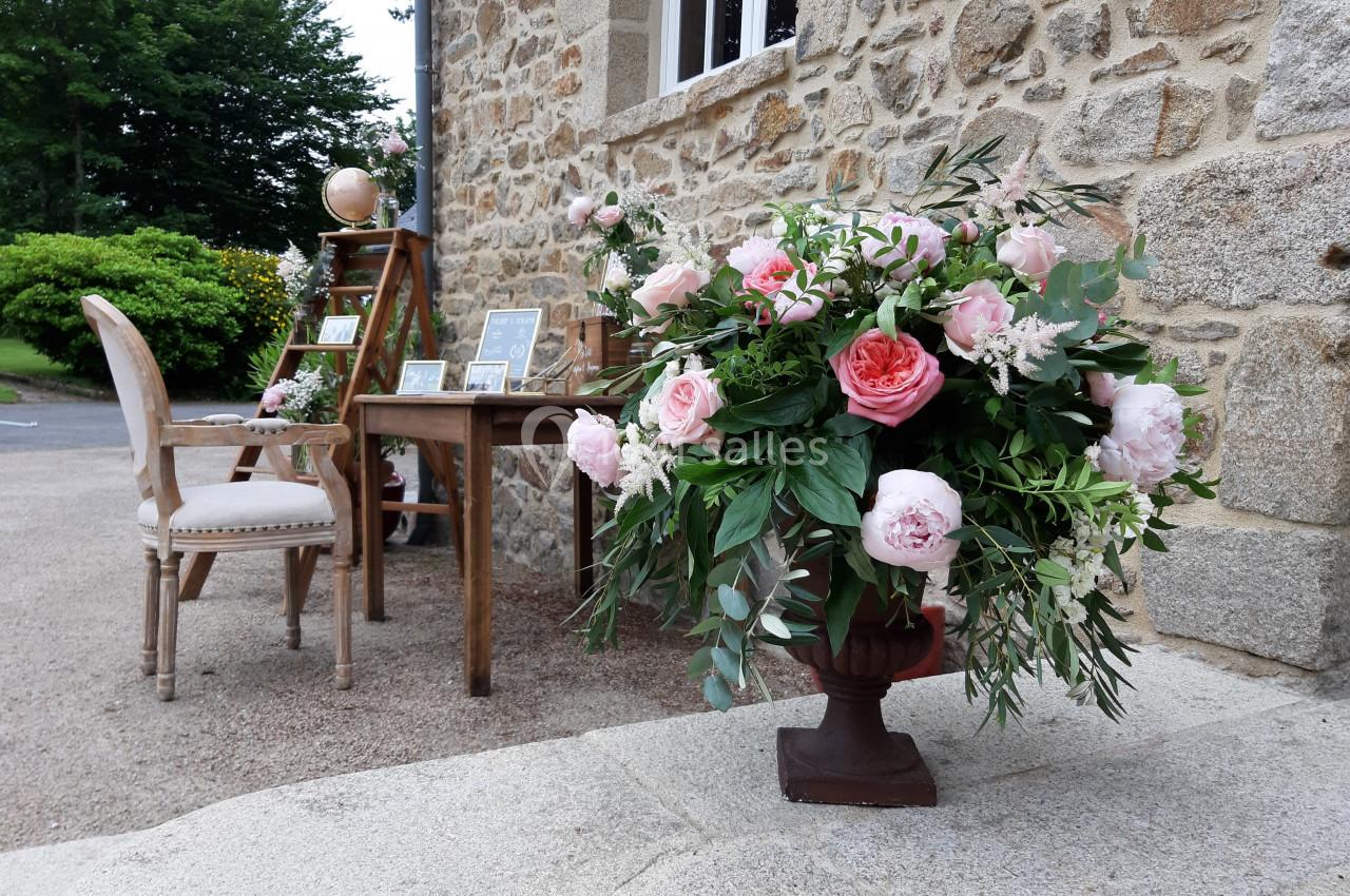 Arrangement floral avec des roses roses et feuillage devant une table en bois et une façade en pierre.