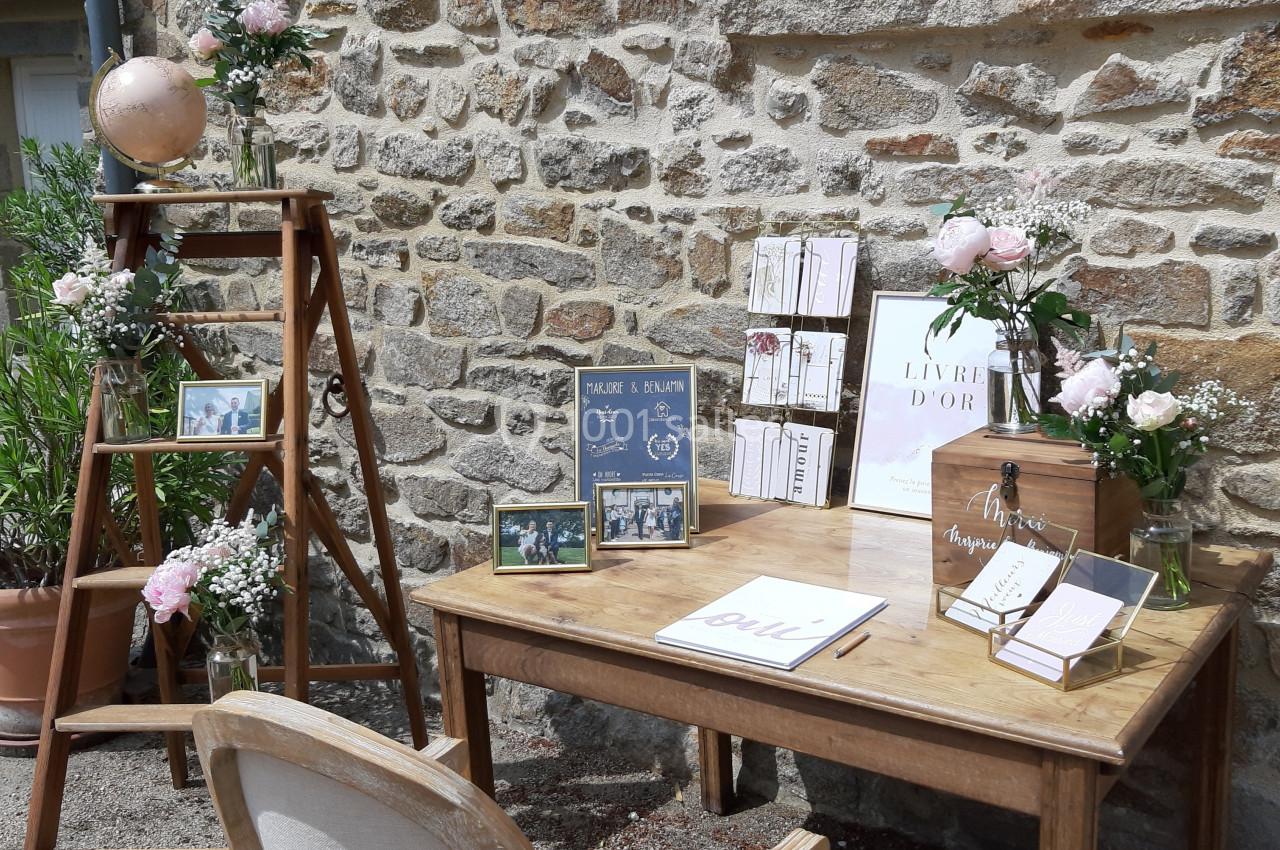 Table en bois avec livre d'or, décorations florales et échelle ornée de cadres, devant un mur en pierre.