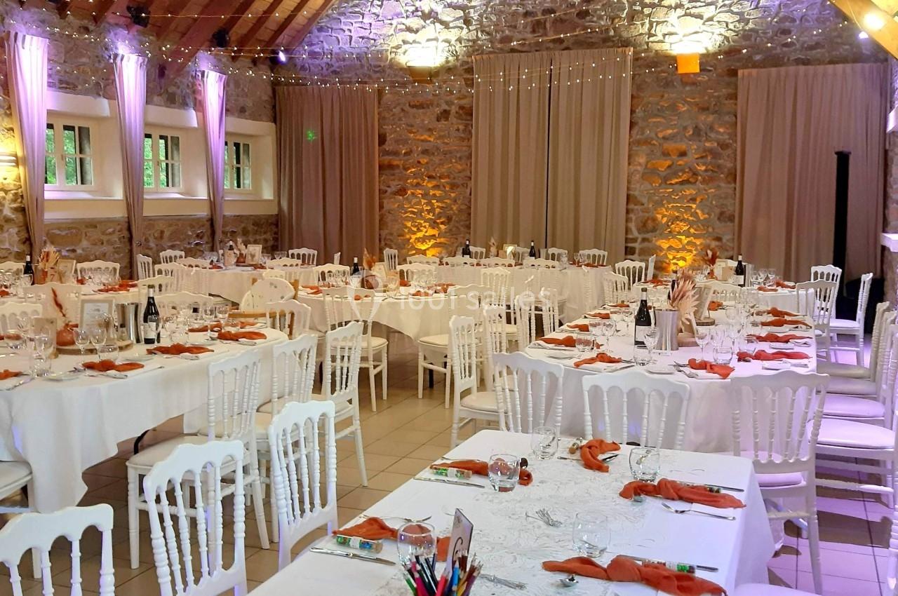 Salle de réception avec tables dressées, nappes blanches, serviettes orange, chaises blanches et éclairage chaleureux.