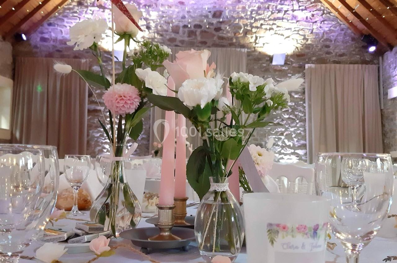 Table de réception décorée avec des fleurs, bougies roses et vaisselle élégante dans une salle en pierre avec poutres…
