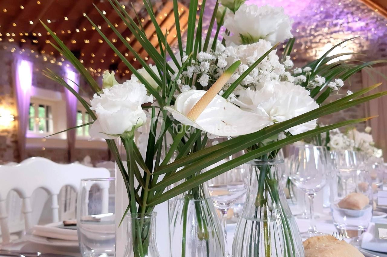 Vases en verre contenant des fleurs blanches et feuillages, disposés sur une table dressée dans une salle avec poutres…