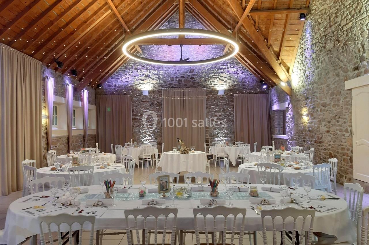 Salle de réception en pierre avec tables décorées, chaises blanches et éclairage chaleureux sous un plafond en bois.