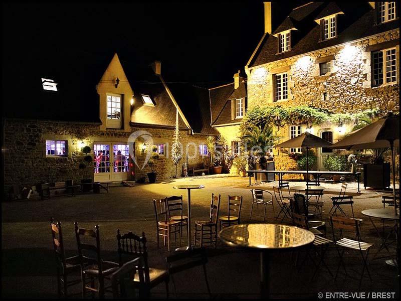 Cour d'une bâtisse en pierre éclairée la nuit, avec des tables et chaises disposées en extérieur.