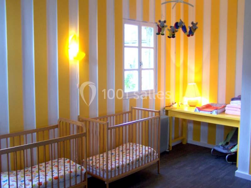 Deux lits pour bébé en bois dans une chambre aux murs rayés jaunes et blancs, avec une table et des luminaires.