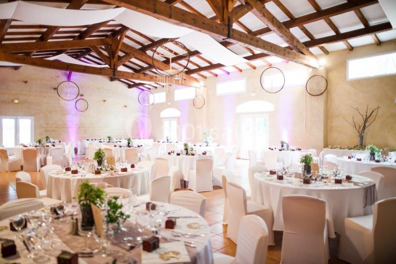 Salle de réception lumineuse avec tables rondes dressées, chaises blanches et poutres apparentes au plafond.