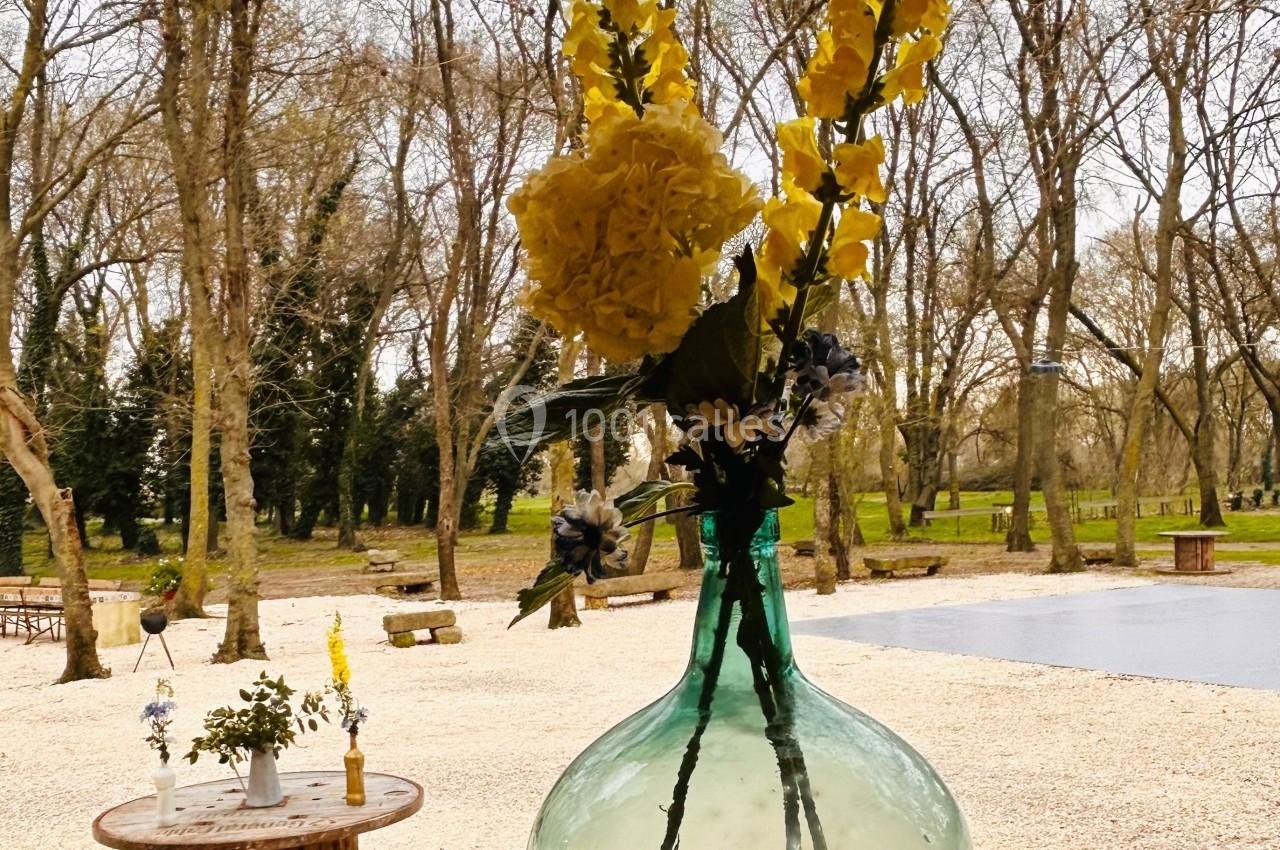 Vase en verre transparent avec des fleurs jaunes et blanches, posé sur une table devant un jardin arboré en hiver.