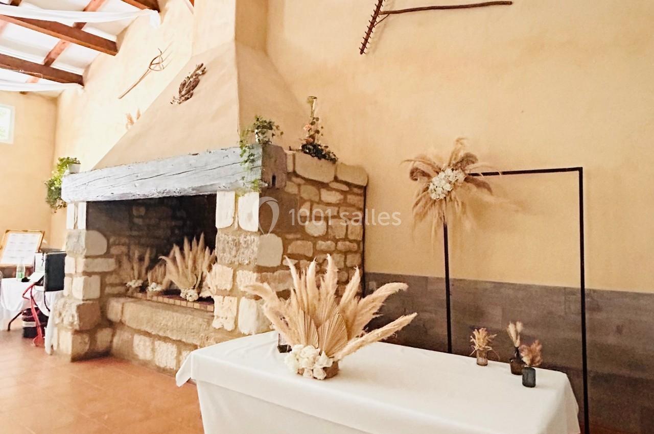 Cheminée en pierre dans une salle rustique avec une table décorée de fleurs séchées et un cadre métallique.